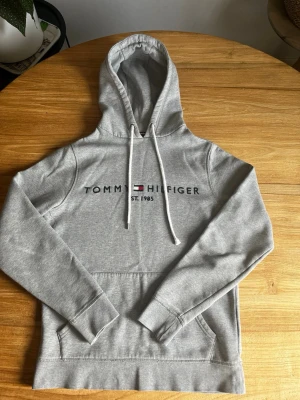 Grå hoodie från Tommy Hilfiger - Snygg grå hoodie från Tommy Hilfiger med klassisk logga och text på bröstet samt liten flagga på ärmen. Hoodien har huva med vita snören och en stor magficka. Tillverkad i mjuk bomullsblandning, perfekt för en chill och stilren look. 