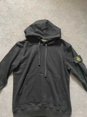 Mörkgrå hoodie med Stone Island patch - Snygg mörkgrå hoodie från Stone Island med klassisk huva och dragsnören. Stor känguruficka framtill och ikonisk avtagbar patch med logga på vänster ärm. Perfekt för en avslappnad streetwear-look.