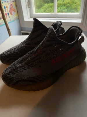 Grå Yeezy Boost 350 V2 sneakers - Säljer ett par Yeezy Boost 350 V2 sneakers i grått med mönstrad ovandel och röd text på sidan. Skorna har en chunky sula och är tillverkade i stickat material för en skön och flexibel känsla. Perfekta för dig som gillar streetwear och vill sticka ut.