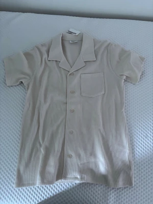 Beige skjorta från Kappahl - Kortärmad beige skjorta från Kappahl med klassisk krage och bröstficka. Skjortan har knappar framtill och är tillverkad i ett strukturerat bomullstyg som ger en avslappnad känsla. Perfekt för varma dagar.