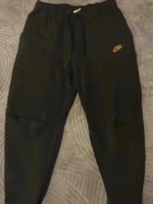 Svarta Nike mjukisbyxor med logga - Säljer ett par svarta mjukisbyxor från Nike med guldfärgad logga på vänster lår. Byxorna har dragsko i midjan, ribbade muddar vid bensluten och fickor på sidorna. Perfekta för chill dagar eller träning.