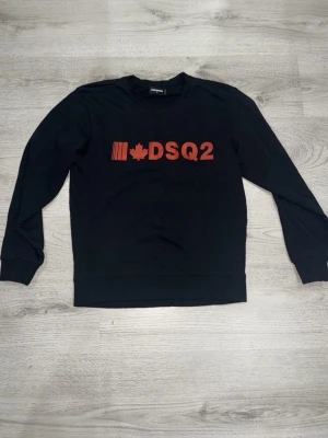 Svart sweatshirt från Dsquared2 - Svart sweatshirt från Dsquared2 med röd DSQ2-logga och lönnlöv på bröstet. Tröjan har rund halsringning, långa ärmar och ribbade muddar vid ärmslut och nederkant. Tillverkad i mjukt bomullsmaterial för en skön och avslappnad look.