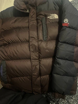 Brun dunjacka The North Face - Säljer en brun dunjacka från The North Face Summit Series med svarta detaljer. Jackan har hög krage, dragkedja, bröstficka och sidofickor. Perfekt för kalla vinterdagar och riktigt varm tack vare dunfyllningen.
