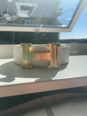 Vitt bälte med stor guldfärgad H-spänne - Snyggt vitt bälte med ett stort, guldfärgat H-format spänne. Bältet har en stilren och modern look som passar till många outfits. Tillverkat i skinn för en exklusiv känsla och hållbarhet.