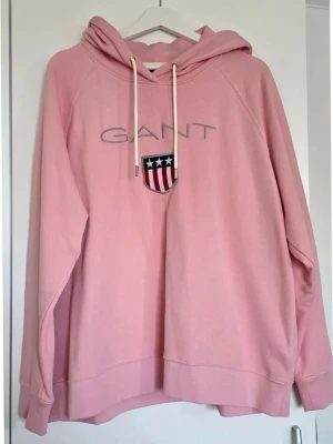 Rosa hoodie från GANT  - Säljer en ljusrosa hoodie från GANT med broderad logga och sköld på bröstet. Hoodien har huva med vita dragsnören och avslappnad passform. Perfekt för en chill och snygg look. Materialet känns mjukt och bekvämt.