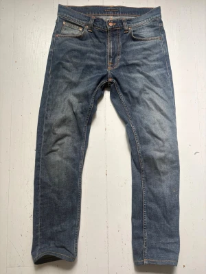 Nudie Jeans Lean Dean - Klassiska blå jeans med rak passform och snygga orange kontrastsömmar. Jeansen har fem fickor och subtila slitningar på bakfickorna. Tillverkade i denim med en autentisk tvättad look, perfekta för en avslappnad stil. Storlek 29/30