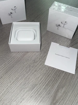 AirPods gen 2 - Säljer ett par vita trådlösa hörlurar från Apple med matchande laddningsetui. Hörlurarna har en stilren och kompakt design i blank plast och levereras i originalförpackning. Perfekta för musik, samtal och träning.