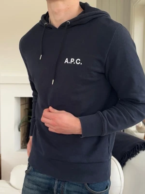 Mörkblå hoodie från A.P.C. - Snygg mörkblå hoodie från A.P.C. med vit logga på bröstet. Klassisk modell med huva och dragsko samt ribbade muddar vid ärmslut och midja. Perfekt för en avslappnad och stilren look.