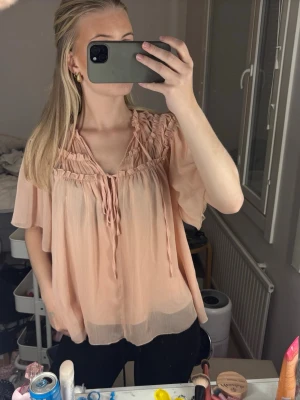 Ljusrosa luftig blus med volang - Söt ljusrosa blus i ett tunt, transparent tyg med volangdetaljer vid axlarna och knytband vid halsen. Blusen har en lös passform och korta, fladdriga ärmar som ger en romantisk vibe. Perfekt att styla med jeans eller kjol för en trendig look.