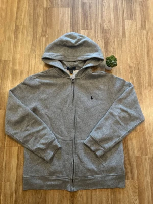 Grå zip hoodie från Polo Ralph Lauren - Snygg grå hoodie med dragkedja från Polo Ralph Lauren. Klassisk design med huva, ribbade muddar och fickor framtill. Liten broderad logga på bröstet. Perfekt för en avslappnad och stilren look. Helt ny