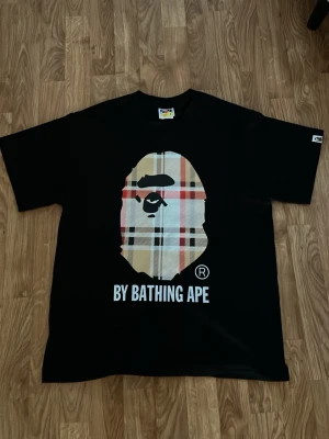 Svart BAPE t-shirt med logga - Svart t-shirt med stort BAPE-tryck framtill i beige och röd rutig design samt texten 'BY BATHING APE' under. Klassisk passform och rund halsringning. Perfekt för dig som gillar streetwear och vill sticka ut.
