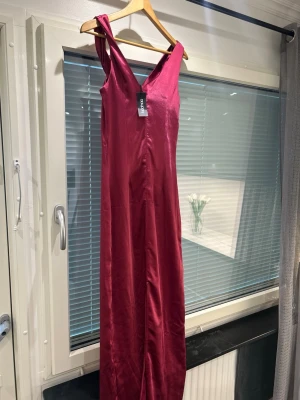Rosa/lila  långklänning i satin - Elegant rosa/lila långklänning i glansigt satinliknande material. Klänningen har breda axelband och en snygg draperad urringning framtill. Passformen är rak och smickrande, perfekt för speciella tillfällen.
