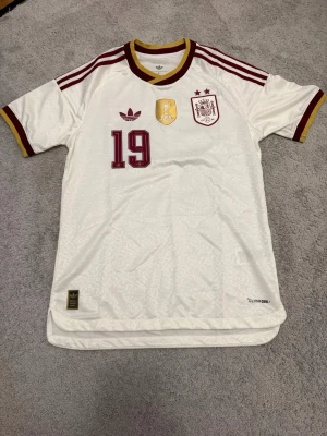 Spaniens matchtröja Adidas nr 19 - Säljer en vit matchtröja från Spaniens landslag med vinröda detaljer och nummer 19. Tröjan har Adidas-logga, FIFA-märke och landslagsemblem på bröstet. Rund hals med vinröd och gul kant, samt klassiska Adidas-ränder på axlarna. Tillverkad i lätt funktionsmaterial.
