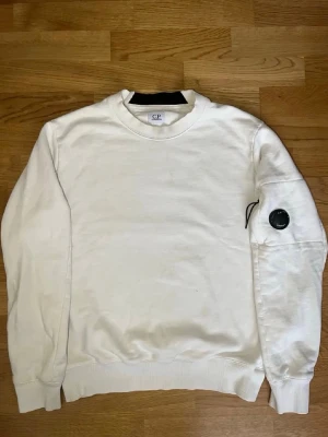 Vit sweatshirt från C.P. Company - Vit sweatshirt från C.P. Company med rund halsringning och ribbade muddar. Tröjan har en klassisk passform och en svart detalj på ena ärmen som ger en cool touch. Perfekt för en avslappnad och stilren look.