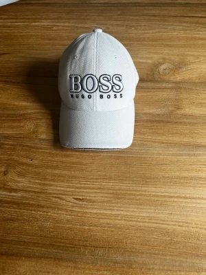 Vit keps från Hugo Boss - Snygg vit keps från Hugo Boss med tydlig svart brodyr på framsidan där det står 'BOSS HUGO BOSS'. Kepsen har klassisk böjd skärm och justerbart band bak. Tillverkad i ett strukturerat tyg som ger en sportig känsla.