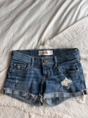 Blå jeansshorts från Hollister - Säljer ett par blå jeansshorts från Hollister med slitna detaljer och råa kanter. Klassisk femficksmodell med broderade bakfickor och liten logga fram. 