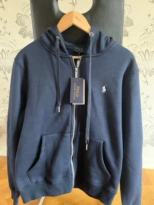 Marinblå hoodie från Polo Ralph Lauren - Snygg marinblå hoodie från Polo Ralph Lauren med dragkedja framtill, två fickor och klassisk huva med dragsko. Ikoniska loggan broderad på bröstet. Perfekt för en avslappnad och stilren look.