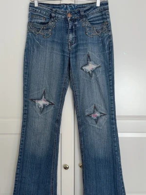 Blå bootcut jeans med stjärnslitage - Säljer ett par blå bootcut jeans som är låg midjade, med slitningar i form av stjärnor på benen och broderade detaljer vid fickorna. Jeansen har en klassisk femficksmodell och utsvängda ben. Materialet är denim i bomull med snygg tvätt och slitningar för en trendig look.