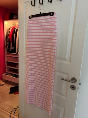 Rosa och vit randig långkjol - en långkjol med raka linjer i ett stickat material. Kjolen har ett randigt mönster i rosa och vitt och är figurnära i modellen. Aldrig använd. För lång för mig som är ca 160cm. 