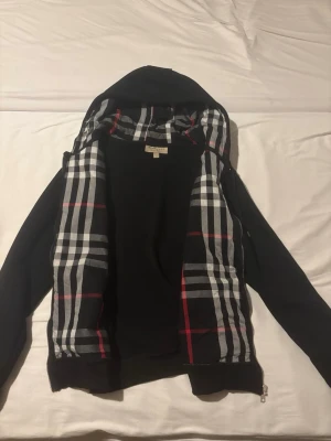 Svart hoodie från Burberry med rutigt foder - Snygg svart hoodie från Burberry med klassiskt rutigt foder i svart, vitt och rött. Dragkedja framtill och huva. Perfekt för dig som vill ha en stilren men ändå lyxig look. Materialet känns mjukt och bekvämt, och detaljerna ger en exklusiv känsla.