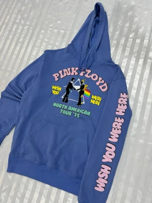 Blå Pink Floyd hoodie med tryck - Blå hoodie med stor huva och magficka. Framsidan har färgglatt Pink Floyd-tryck med texten 'Wish You Were Here' och 'North American Tour '75'. På ena ärmen står det 'Wish You Were Here' i stora rosa bokstäver. Perfekt för dig som gillar bandtröjor och retrostil.