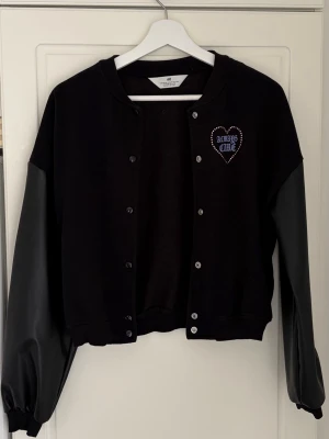 Svart bomberjacka med tryck, från H&M, 158 / 164 - Snygg svart bomberjacka med texten 'ALWAYS CARE' i lila på bröstet och ryggen, inramad av hjärtan. Jackan har knäppning framtill och kontrasterande ärmar i ett glansigt material. Perfekt för en trendig streetwear-look.