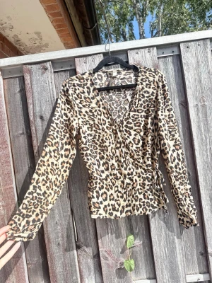 Leopardmönstrad omlottblus - Snygg leopardmönstrad omlottblus med knyt i midjan och v-ringning. Blusen har långa ärmar och är tillverkad i ett lätt, mjukt tyg som ger en skön passform. Perfekt för dig som vill sticka ut med ett trendigt djurmönster.