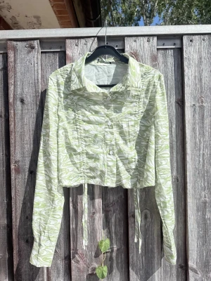 Grönvit mönstrad croppad blus oanvänd - Trendig croppad blus i grönvitt swirl-mönster med långa ärmar och knappar framtill. Blusen har snörning i midjan för justerbar passform och en klassisk krage. Perfekt för dig som vill sticka ut med en cool och unik look.