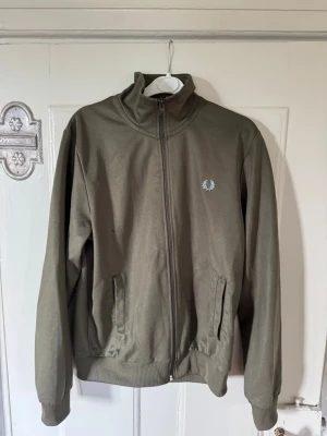 Olivgrön zipjacka från Fred Perry - Snygg olivgrön ziptröja från Fred Perry med hög krage och ribbade muddar. Tröjan har två fickor framtill och den klassiska Fred Perry-loggan broderad på bröstet. Perfekt för en avslappnad och sportig stil. Finns en liten svart prick på bröstet annars i fint skick.