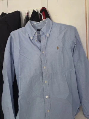 Ljusblå Ralph lauren skjorta - Klassisk ljusblå skjorta med button-down krage och broderad ralp lauren logga på bröstet. Skjortan har långa ärmar och vita knappar framtill. Perfekt till jeans eller chinos för en clean look.