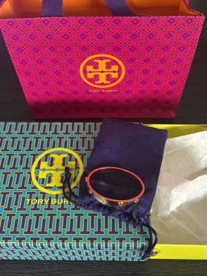 Guldigt armband från Tory Burch - Snyggt armband från Tory Burch i guldfärgad metall. Armbandet har små logodetaljer runt om och en stilren, modern design. Perfekt accessoar för att lyfta din outfit. Levereras med originalpåse och ask.