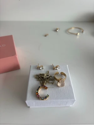 Caroline Svedbom smycken  - Säljer ett set med guldfärgade smycken: halsband med C-berlock dekorerad med färgglada stenar, örhängen med klara stenar, ring med stor ljus sten och ett armband i bakgrunden. Perfekt för dig som gillar att mixa och matcha accessoarer.