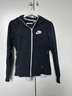 Svart Nike zip hoodie med huva - Snygg svart hoodie från Nike med dragkedja framtill och vit logga på bröstet. Tröjan har huva och vita detaljer längs dragkedjan och insidan av kragen. Perfekt för en sportig och avslappnad stil.