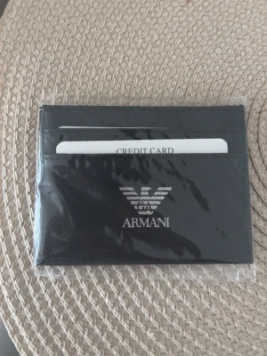 Svart kortfodral från Armani - Snyggt svart kortfodral från Armani med silverfärgad logga framtill. Tillverkat i slätt läder och har flera fack för kort. Perfekt för dig som vill ha en stilren och smidig plånbok att bära med sig.
