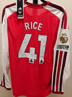 Arsenal långärmad hemma 26/27 spelar version  Rice 41 - Snygg långärmad Arsenal matchtröja från Adidas med Rice 41 på ryggen. Tröjan är röd med vita och vinröda detaljer samt klubbmärke och sponsortryck framtill. Tillverkad i ventilerande funktionsmaterial, perfekt för fotboll eller träning.