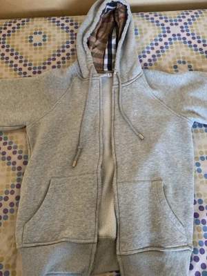Burberry zip up - Snygg grå hoodie från Burberry med dragkedja, fickor framtill och klassiskt rutigt foder i huvan. Broderad logga på bröstet och ribbade muddar. Perfekt för en chill och stilren look.