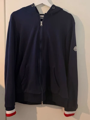 Moncler zip  - Snygg marinblå hoodie från Moncler med dragkedja framtill och huva. Rödvita detaljer på ärmsluten och luvan. Moncler-logga på vänster ärm. Kan eventuellt vara öppen för byten!