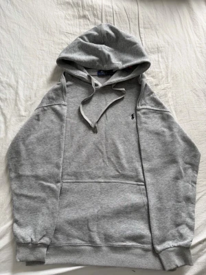 Grå hoodie från Polo Ralph Lauren - Klassisk grå hoodie från Polo Ralph Lauren med broderad logga på bröstet. Hoodien har dragsko i huvan, stor magficka och ribbade muddar vid ärmar och nederkant. Tillverkad i mjukt sweatshirtmaterial som är perfekt för chill dagar.