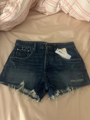 Mörkblå denimshorts med fransig kant - Säljer ett par mörkblå denimshorts med hög midja och rå, fransig kant. Shortsen har klassisk femficksdesign och bälteshällor. Perfekta för sommaren och ger en avslappnad vibe till din outfit.