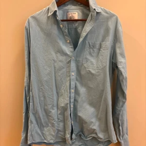Ljusblå skjorta från The Shirt Factory - Säljer en ljusblå skjorta från The Shirt Factory i slim fit-modell. Skjortan har klassisk krage, bröstficka och knappar framtill. Tillverkad i mjuk bomull och passar perfekt till en clean och stilren look.
