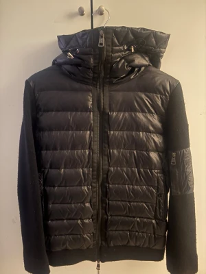 Svart dunjacka från Moncler med huva - Säljer en svart dunjacka från Moncler med huva och dragkedja framtill. Jackan har quiltade partier, mjuka stickade ärmar och en ficka med dragkedja på ena ärmen. Klassisk Moncler-logga på ärmen. Perfekt nu till hösten! Bara att skriva om du har funderingar