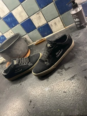 Svarta Saint Laurent sneakers - Pris diskuerbart kom med bud📦 Svarta sneakers från Saint Laurent med vit sula och diskret logga på sidan. Skorna har klassisk låg profil, svarta skosnören och ovandel i tyg. Perfekta för en clean och stilren look. Et litet hål i skon
