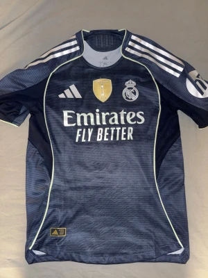 Real Madrid bortatröja Bellingham #5 - Snygg mörkblå Real Madrid bortatröja från Adidas med Bellingham och nummer 5 på ryggen. Tröjan har vita detaljer, klubbmärke och sponsortryck framtill samt Champions League-märke på ärmen. Tillverkad i lätt och ventilerande material, perfekt för fotboll.