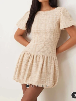 Beige kort klänning med puffärm - Supersöt beige kortklänning med puffiga kortärmar och rund hals. Klänningen har en strukturerad yta och en volangliknande kjol som ger en lekfull vibe. Perfekt för dig som gillar en feminin och trendig stil.