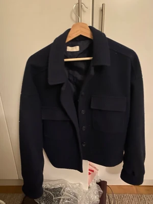 Mörkblå kort kappa från LeGer - Stilren mörkblå overshirt från LeGer med klassisk krage, stora bröstfickor och knäppning framtill. Overshirten har en rak passform och är tillverkad i ett tjockare tyg som ger en snygg och modern look. Perfekt att bära över en hoodie eller t-shirt.