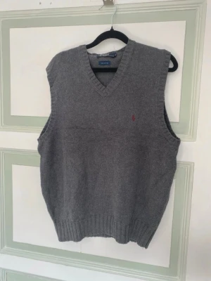 Grå stickad väst från Polo Ralph Lauren - Klassisk grå stickad väst från Polo Ralph Lauren i 100% bomull. Västen har v-ringning och ribbade kanter samt en liten broderad logga på bröstet. Perfekt att slänga över en skjorta för en clean och stilren look.