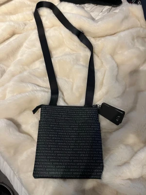 Svart axelväska från Armani Exchange - Snygg svart axelväska från Armani Exchange med justerbar axelrem. Väskan har ett mönster med loggan tryckt över hela ytan och en dragkedja upptill. Perfekt storlek för det viktigaste och en stilren look.