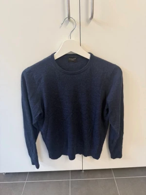 Mörkblå stickad tröja Rose & Born, stl 44/XS - Mörkblå stickad tröja från Rose & Born med rund halsringning och långa ärmar. Tröjan har en klassisk, enkel design och är tillverkad i mjukt material som passar perfekt till lager på lager. Snygg och stilren för en avslappnad look.