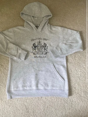 Grå hoodie från Bvaldi med tryck - Snygg ljusgrå hoodie från Bvaldi med stort tryck på bröstet och texten 'Club Bleu Ciel'. Klassisk känguruficka framtill och huva. Tillverkad i mjukt sweatshirtmaterial, perfekt för chill dagar.