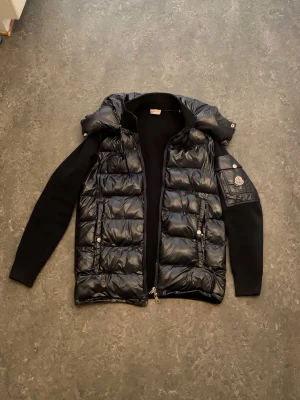 Svart pufferjacka från Moncler - Snygg svart pufferjacka från Moncler med glansig yta och vadderade paneler. Jackan har avtagbar huva, stickade ärmar och två framfickor med tryckknappar. Moncler-logga på ärmen och coolt serietidningsfoder på insidan. Perfekt för kalla vinterdagar.